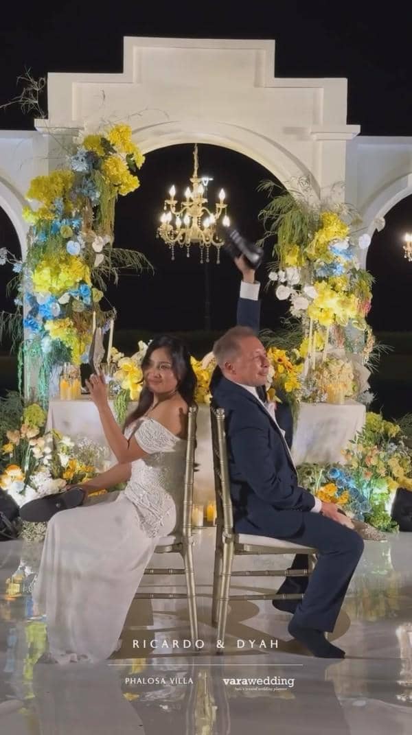 Pasangan pengantin duduk saling membelakangi sambil mengangkat sepatu dalam permainan di resepsi pernikahan dengan dekorasi bunga kuning dan putih.