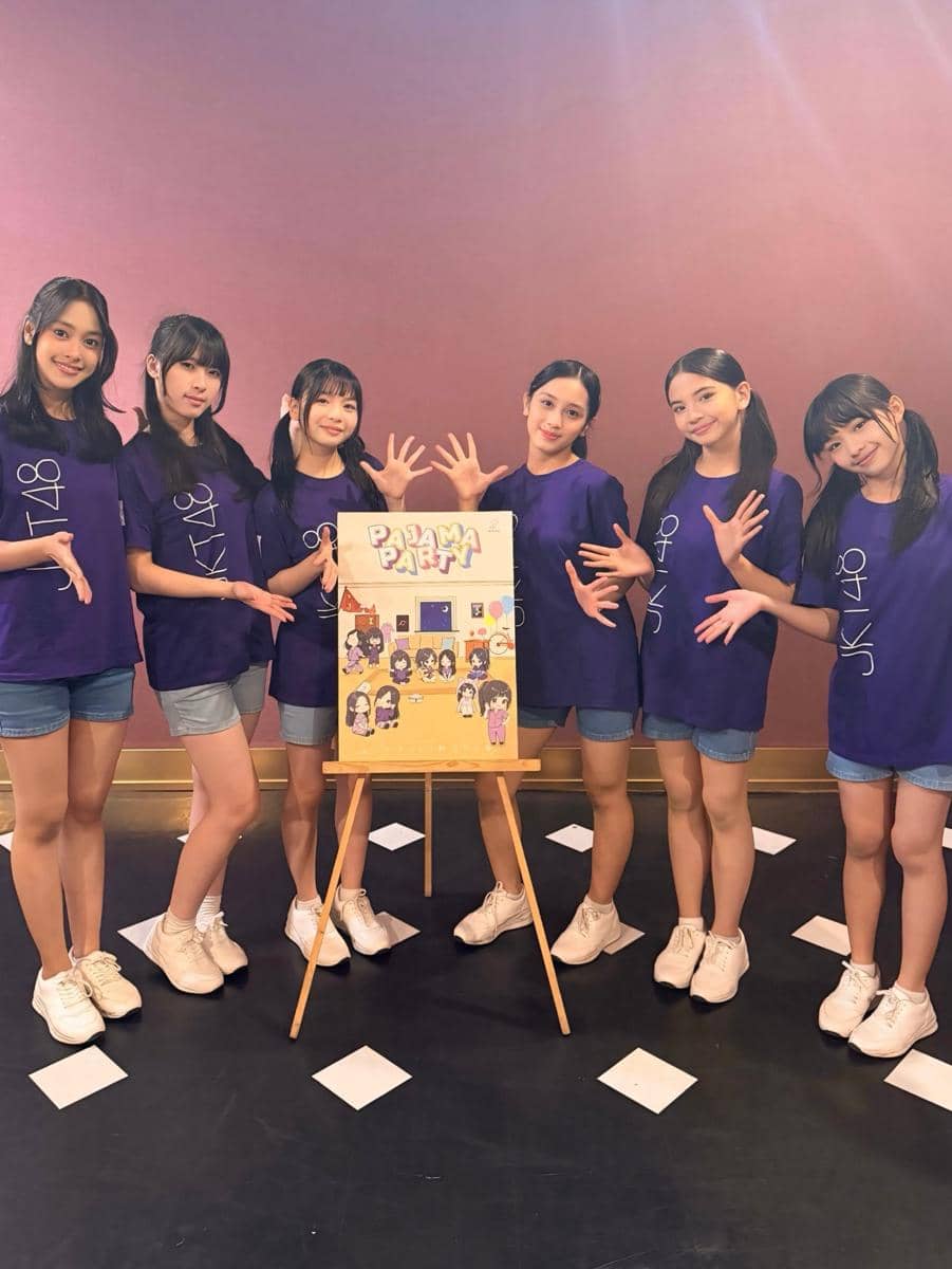 Enam anggota grup idola mengenakan kaus ungu bertuliskan JKT48 berpose bersama papan bertema Pajama Party di ruangan dengan dinding ungu.