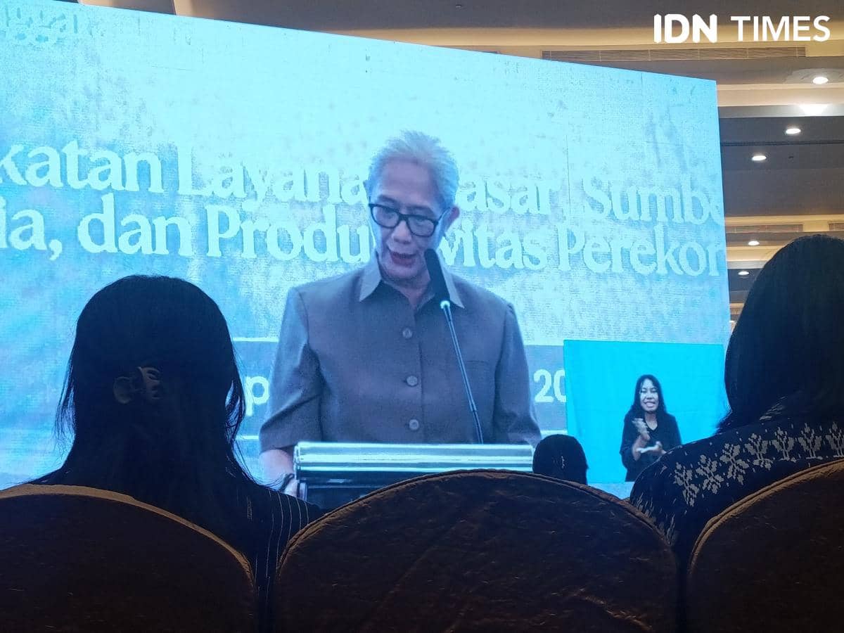 Seorang perempuan berbicara di podium dengan latar layar besar bertuliskan teks presentasi di sebuah acara dalam ruangan.