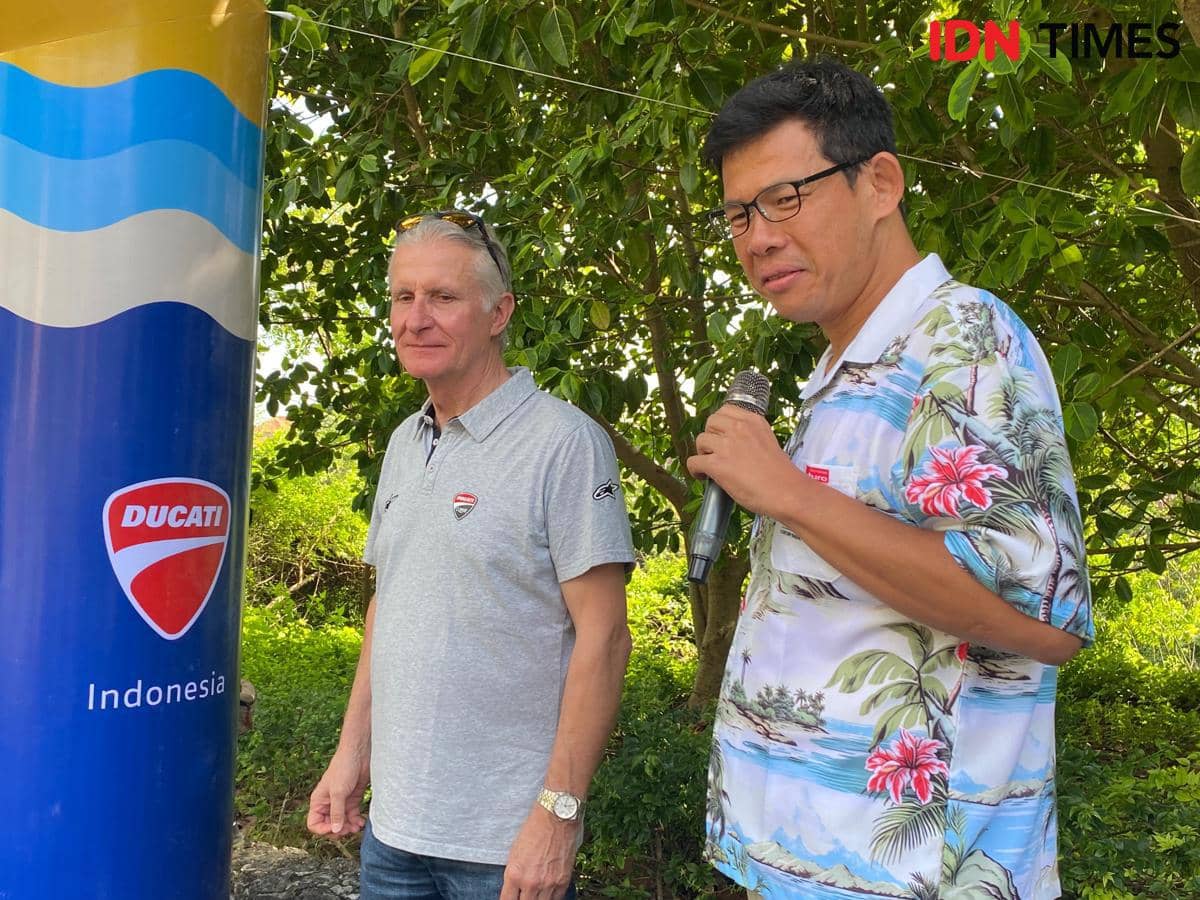 Dua pria berdiri di dekat tiang bertuliskan Ducati Indonesia, salah satunya memegang mikrofon di area luar ruangan dengan latar pepohonan.