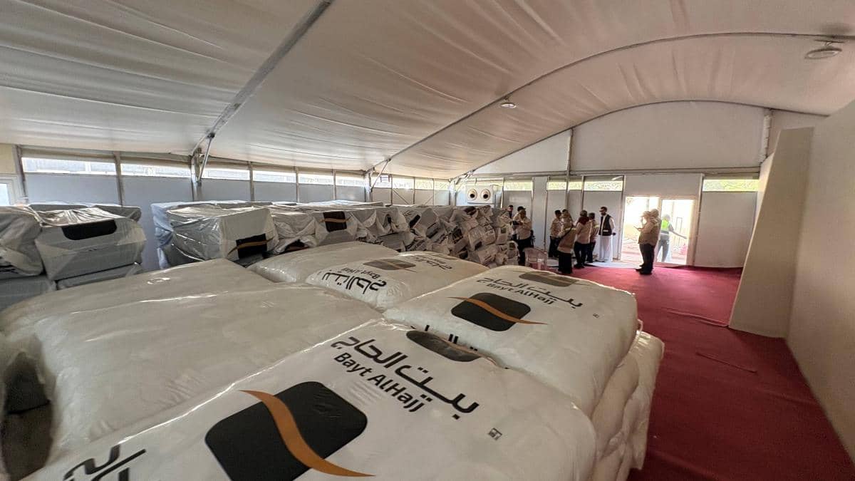 Tumpukan kasur terbungkus plastik bertuliskan Bayt AlHajj tersusun rapi di dalam tenda besar dengan beberapa orang berdiri di pintu keluar.