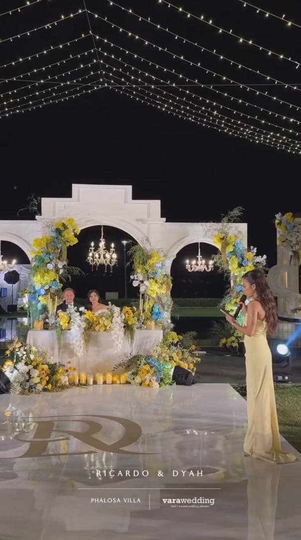 Suasana resepsi pernikahan outdoor dengan dekorasi bunga kuning dan biru, pasangan pengantin duduk di meja utama di bawah lampu gantung.