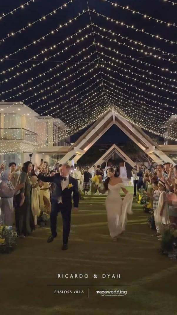 Pasangan pengantin berjalan di bawah hiasan lampu gantung pada resepsi pernikahan malam hari di Phalosa Villa, dikelilingi para tamu yang bersorak.