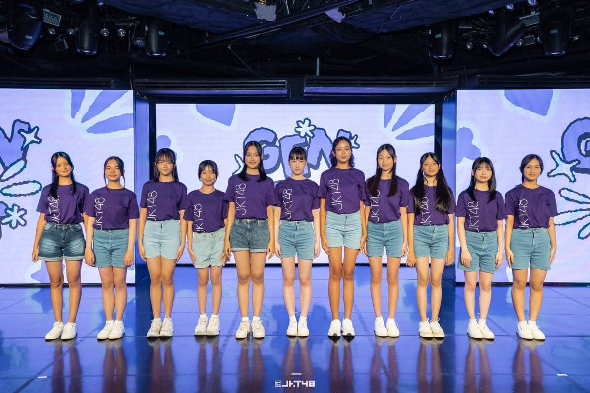 Sebelas anggota grup idola JKT48 berdiri berjajar di atas panggung mengenakan kaus ungu bertuliskan JKT48 dan celana pendek denim.