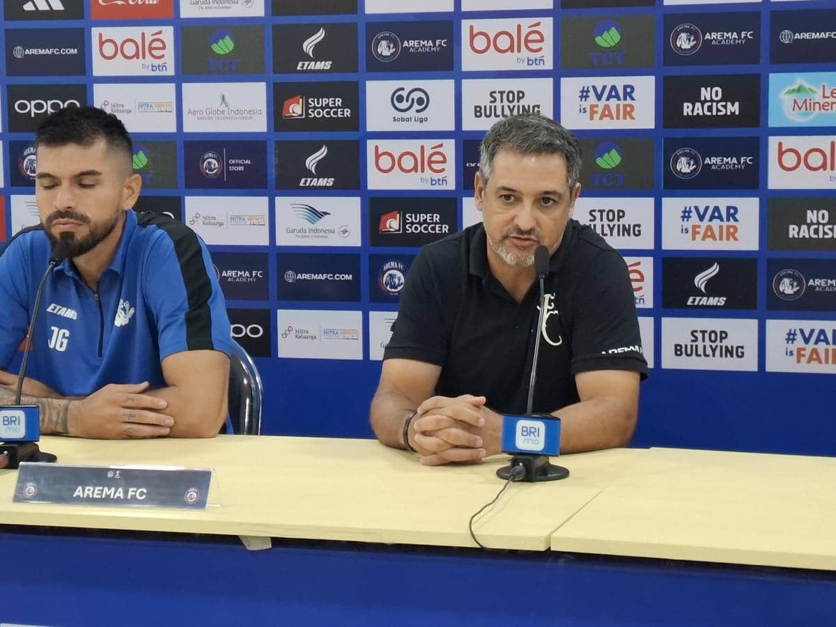Pelatih Arema FC duduk di meja konferensi pers bersama staf tim di depan latar belakang sponsor klub sepak bola.