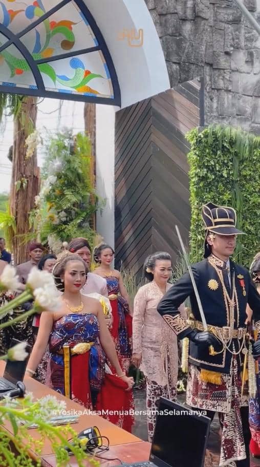Prosesi ngunduh mantu dengan pasangan pengantin mengenakan busana adat Jawa, diikuti keluarga dan tamu di area dekorasi hijau.