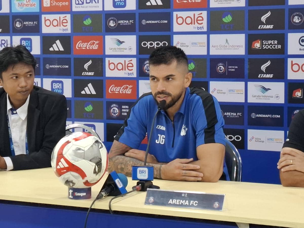 Pemain Arema FC mengenakan seragam biru sedang berbicara di konferensi pers dengan latar belakang logo sponsor klub.