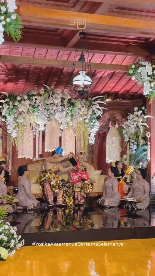 Pasangan pengantin mengenakan busana adat Jawa duduk di pelaminan dengan dekorasi bunga dan ornamen tradisional saat acara ngunduh mantu.