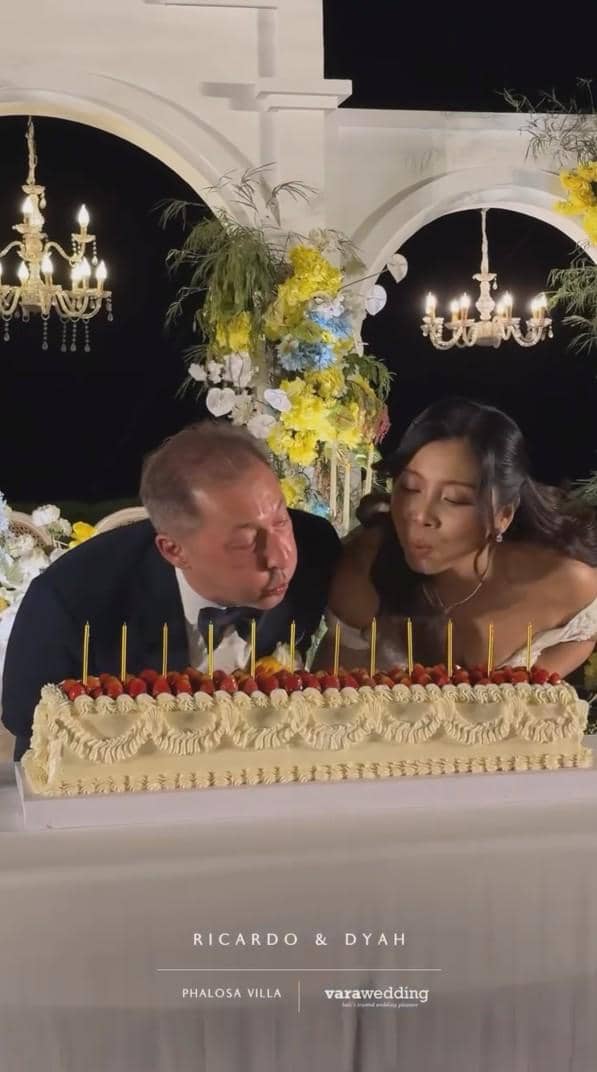 Pasangan pengantin meniup lilin di atas kue pernikahan besar berhias stroberi dalam resepsi malam bertema elegan dengan dekorasi bunga dan lampu gantung.