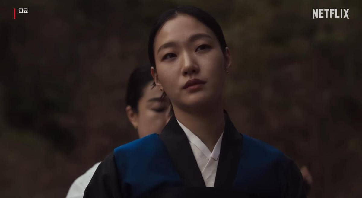 Seorang perempuan mengenakan hanbok berwarna hitam dan biru berdiri dengan ekspresi serius di luar ruangan pada cuplikan film Exhuma.