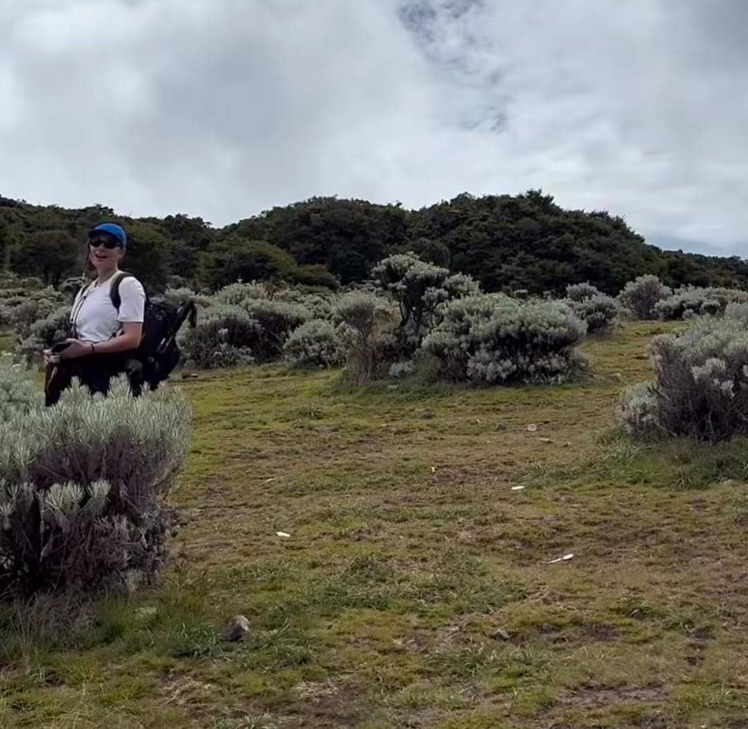Seorang pendaki mengenakan topi biru dan kacamata hitam membawa ransel besar di area sabana Gunung Gede dengan langit berawan.