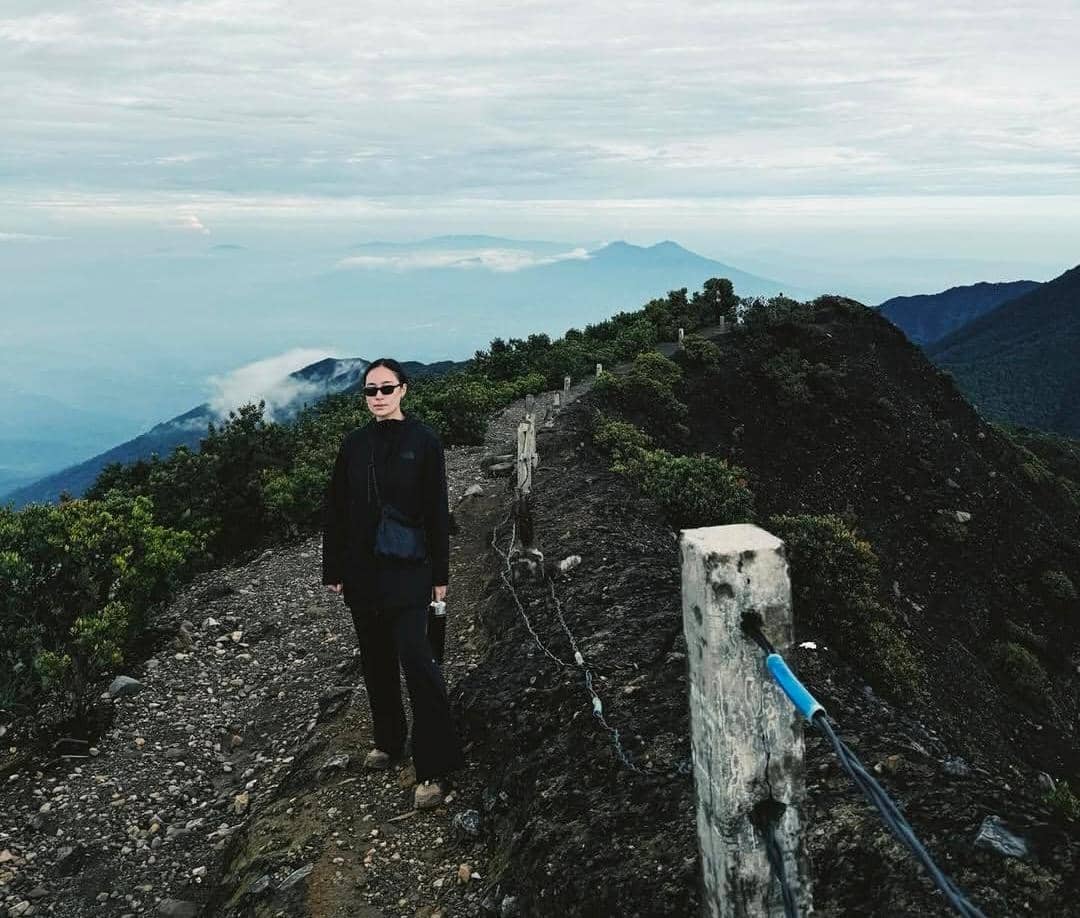 Seorang pendaki mengenakan pakaian hitam berdiri di jalur berbatu Gunung Gede dengan latar pemandangan pegunungan dan awan.