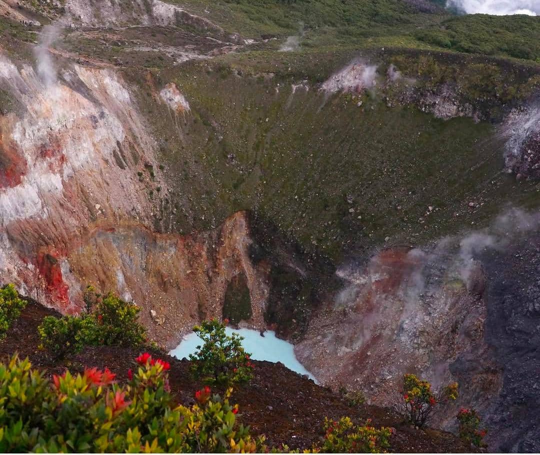 Kawah Gunung Gede dengan dinding curam berwarna cokelat kemerahan, uap panas mengepul, dan air berwarna biru muda di dasar kawah.
