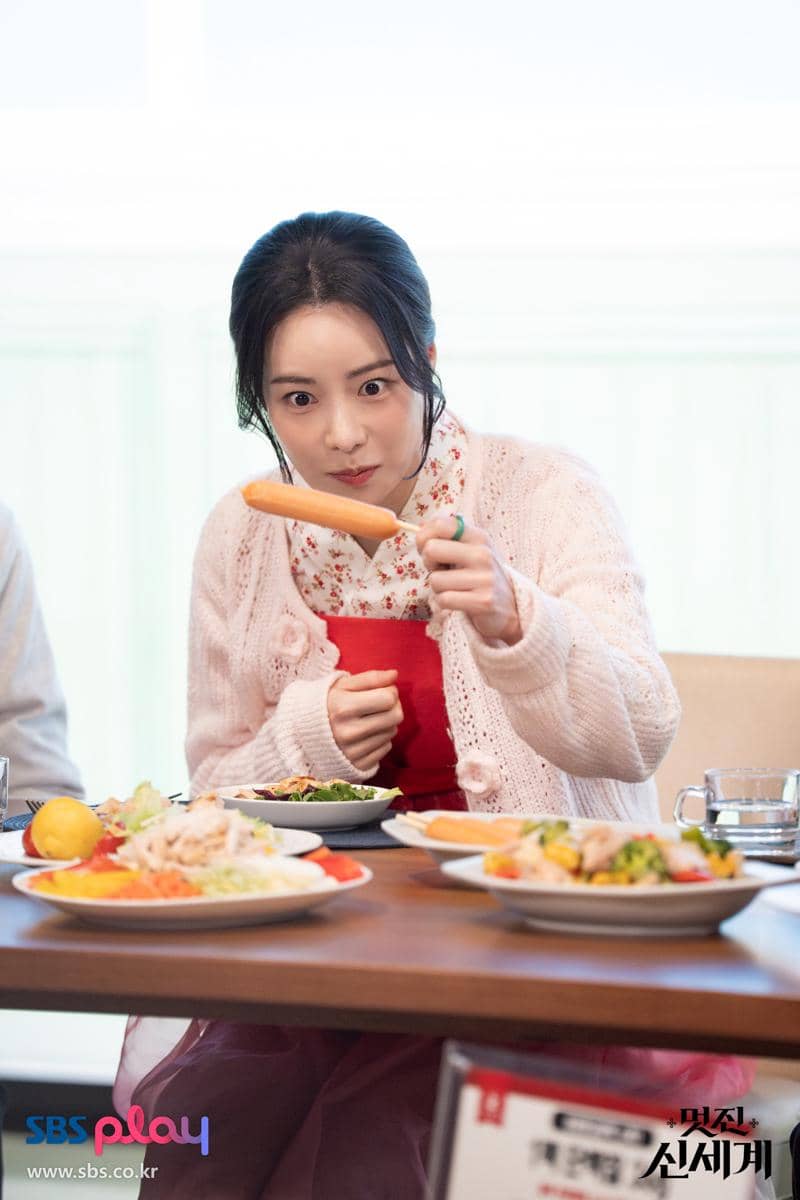 Seorang wanita mengenakan cardigan pink dan apron merah memegang sosis dengan ekspresi terkejut di meja makan penuh hidangan.