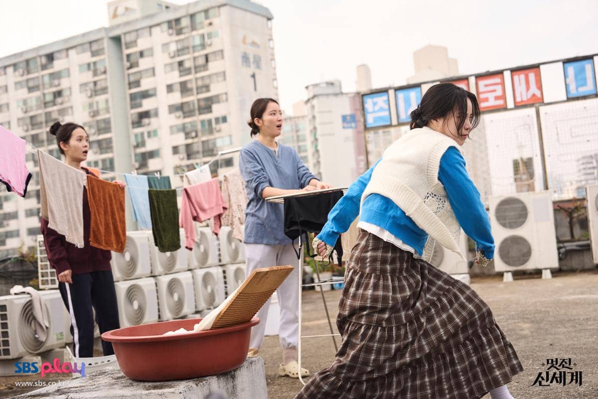 Tiga wanita berada di atap gedung dengan latar apartemen, sedang mencuci dan menjemur pakaian sambil bereaksi terkejut.