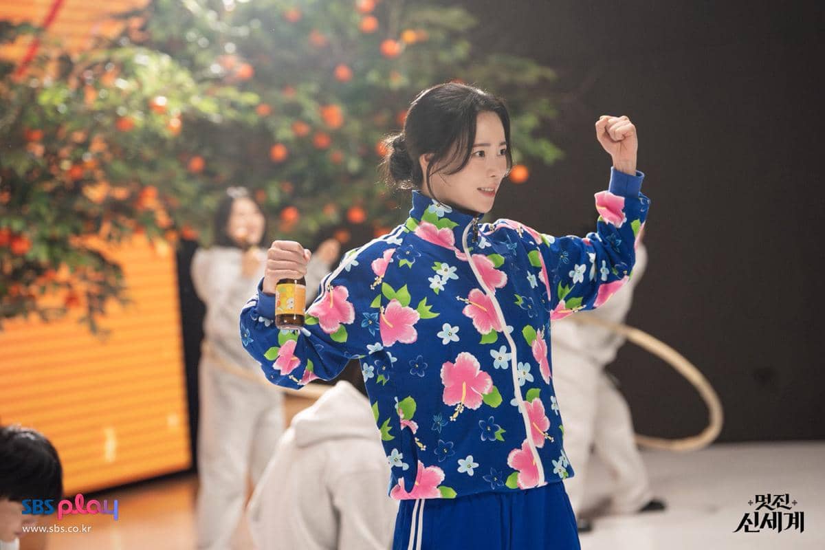 Seorang wanita mengenakan jaket biru bermotif bunga berpose dengan tangan terangkat sambil memegang botol kecil di latar dalam ruangan.