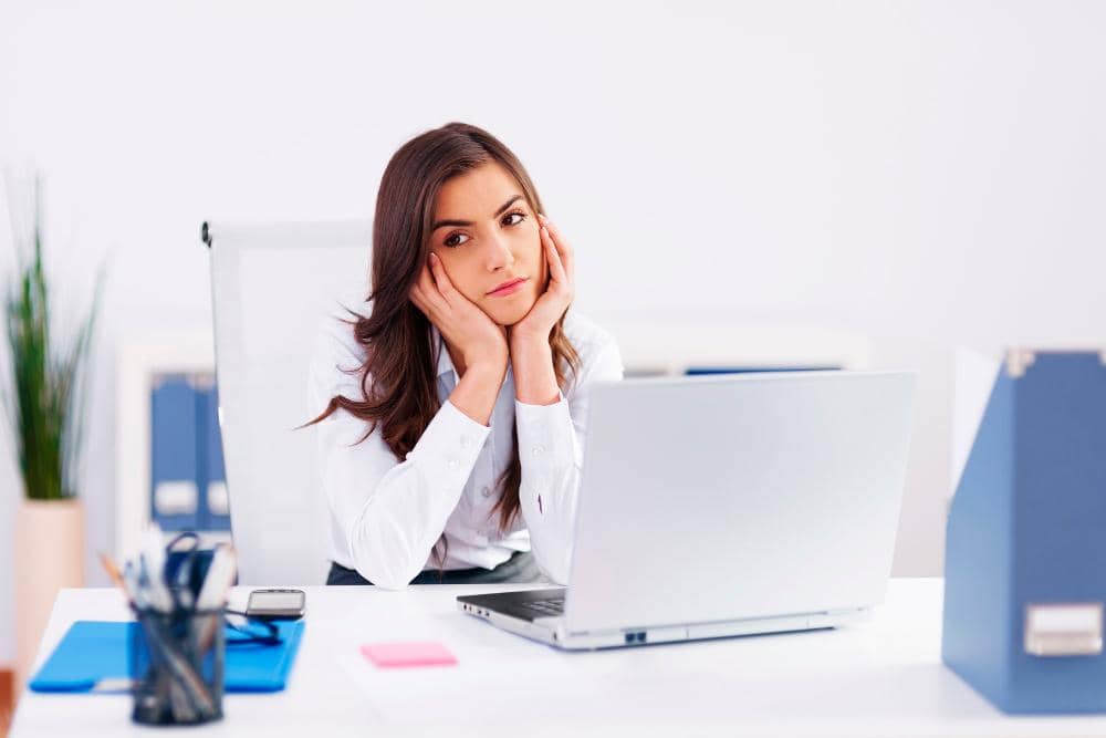 Seorang perempuan duduk di meja kerja dengan tangan menyangga wajah, menatap kosong ke arah laptop di depannya.