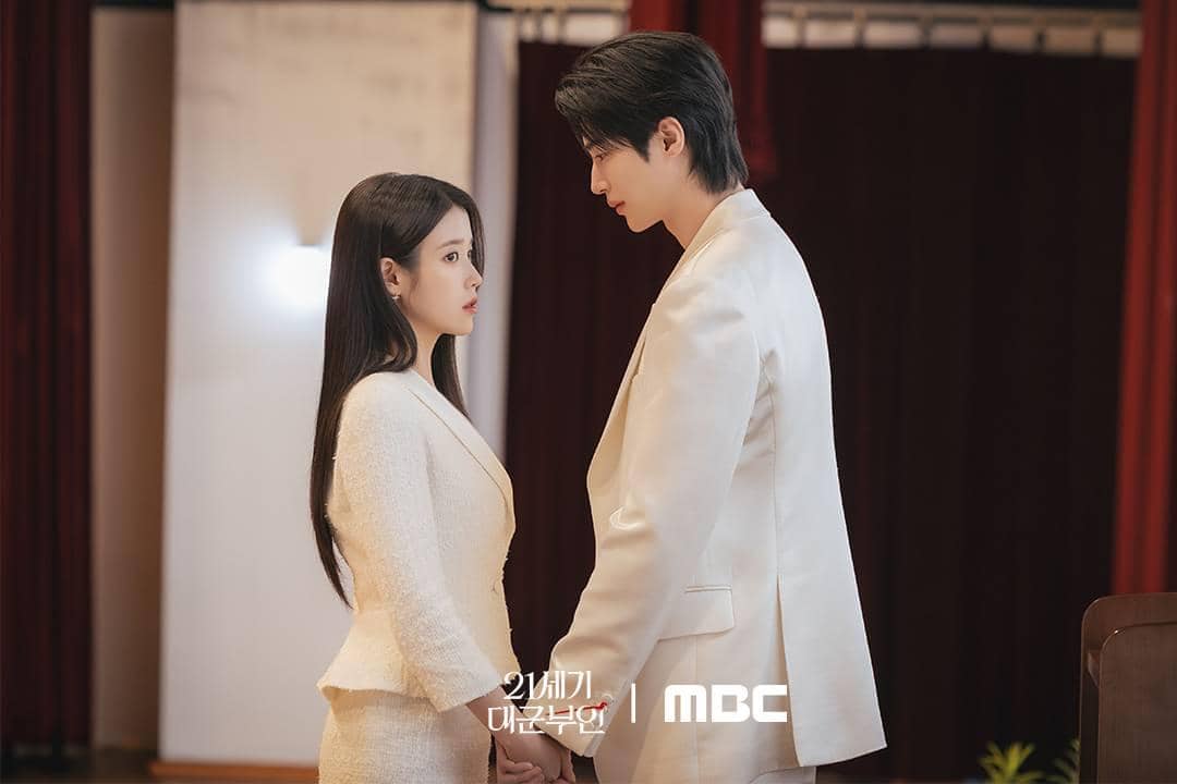 Dua pemeran utama drama Korea Perfect Crown berdiri saling berhadapan dan berpegangan tangan dalam suasana emosional di dalam ruangan.