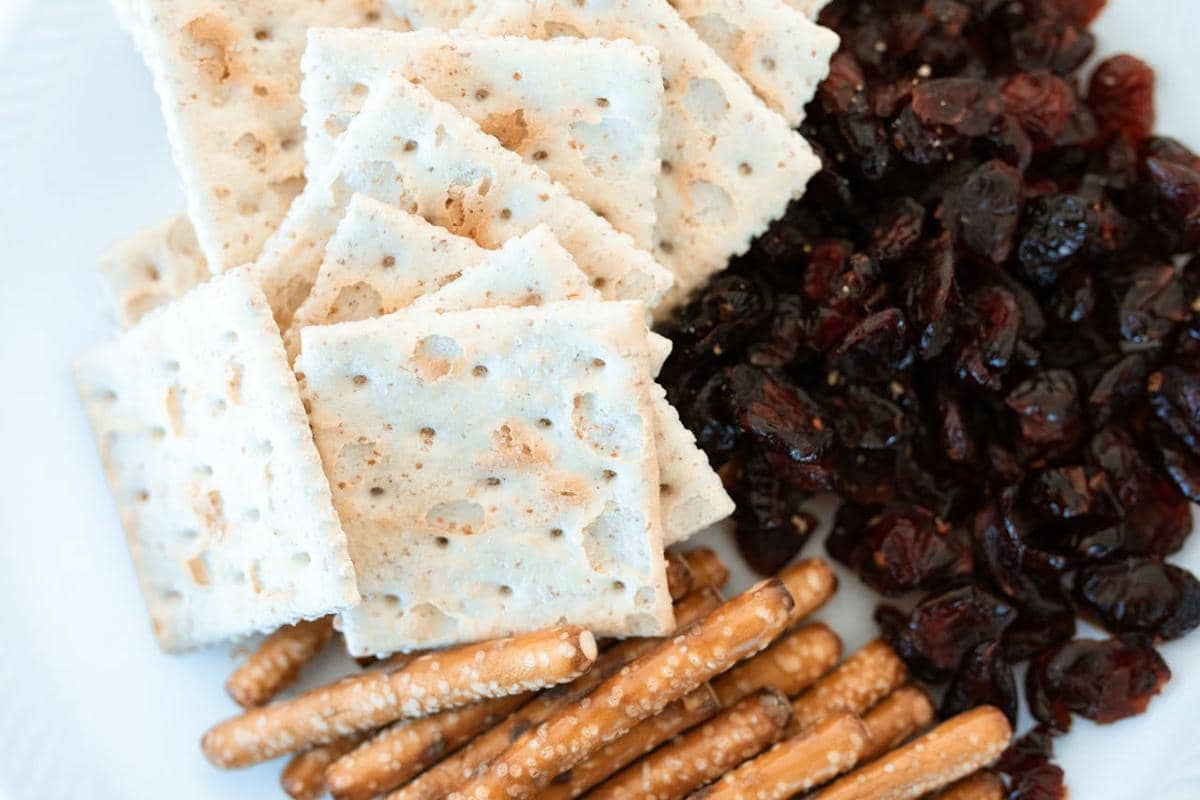 Beberapa potong crackers disajikan bersama pretzel asin dan buah kering berwarna gelap di atas piring putih.