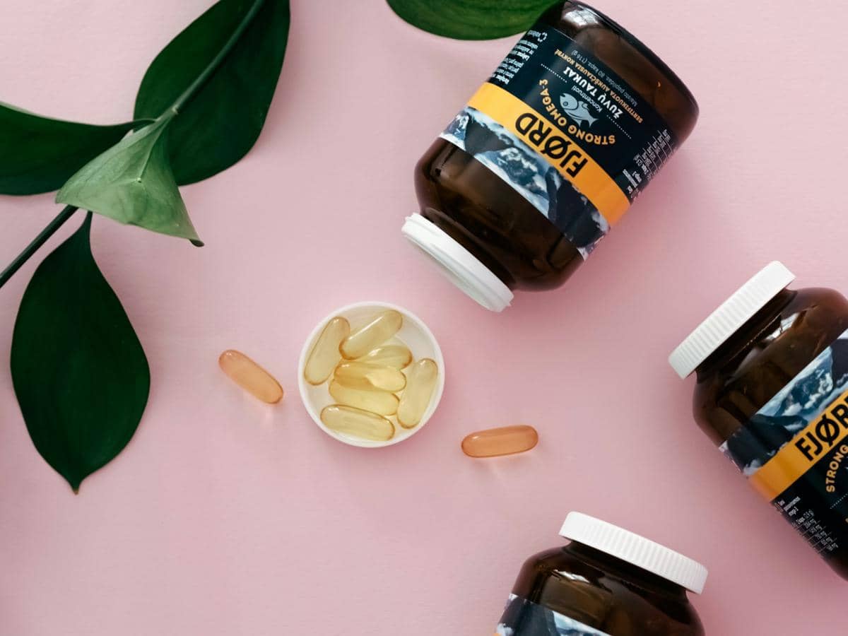 Beberapa botol suplemen omega-3 berwarna cokelat dengan kapsul minyak ikan di atas permukaan berwarna merah muda dan daun hijau di sekitarnya.