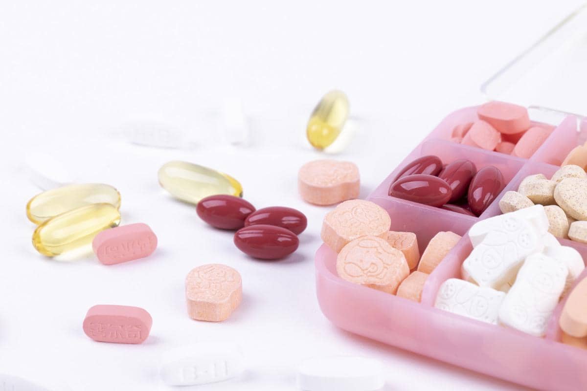 Beragam multivitamin dan suplemen berwarna-warni dalam wadah pil plastik bersekat di atas permukaan putih.
