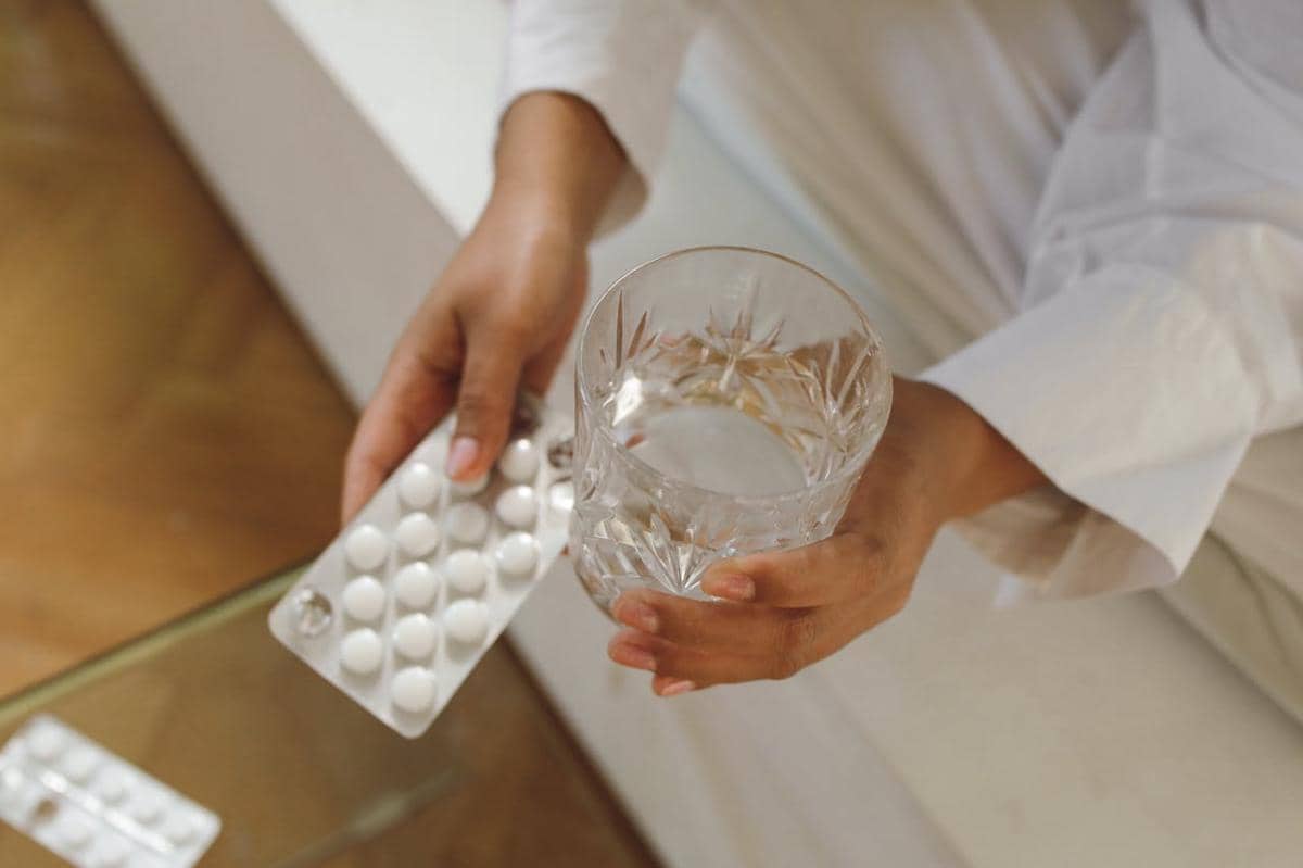 Seseorang memegang segelas air dan blister berisi tablet putih, menggambarkan konsumsi suplemen vitamin B kompleks.