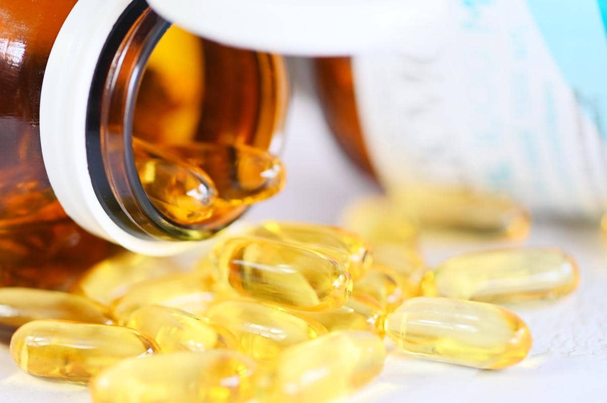 Kapsul vitamin D berwarna kuning keemasan tumpah keluar dari botol cokelat di atas permukaan putih.
