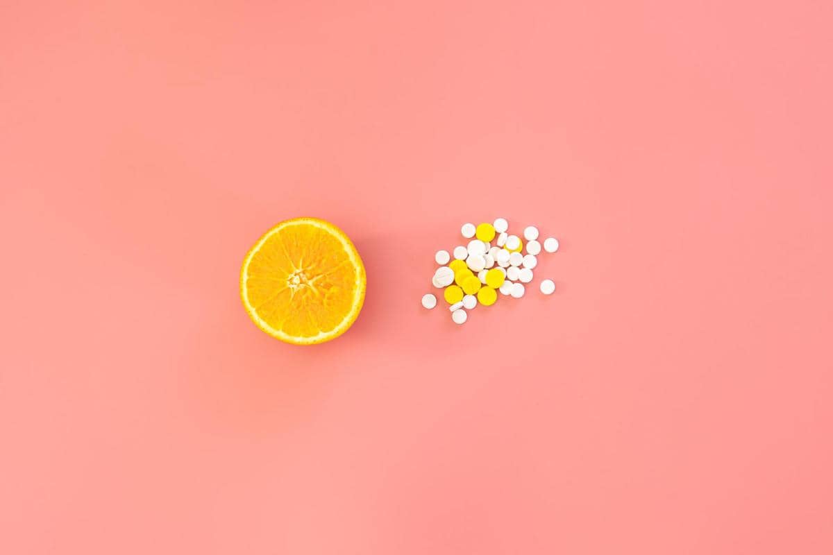 Setengah buah jeruk dan tumpukan tablet vitamin C berwarna putih dan kuning di atas latar belakang merah muda.
