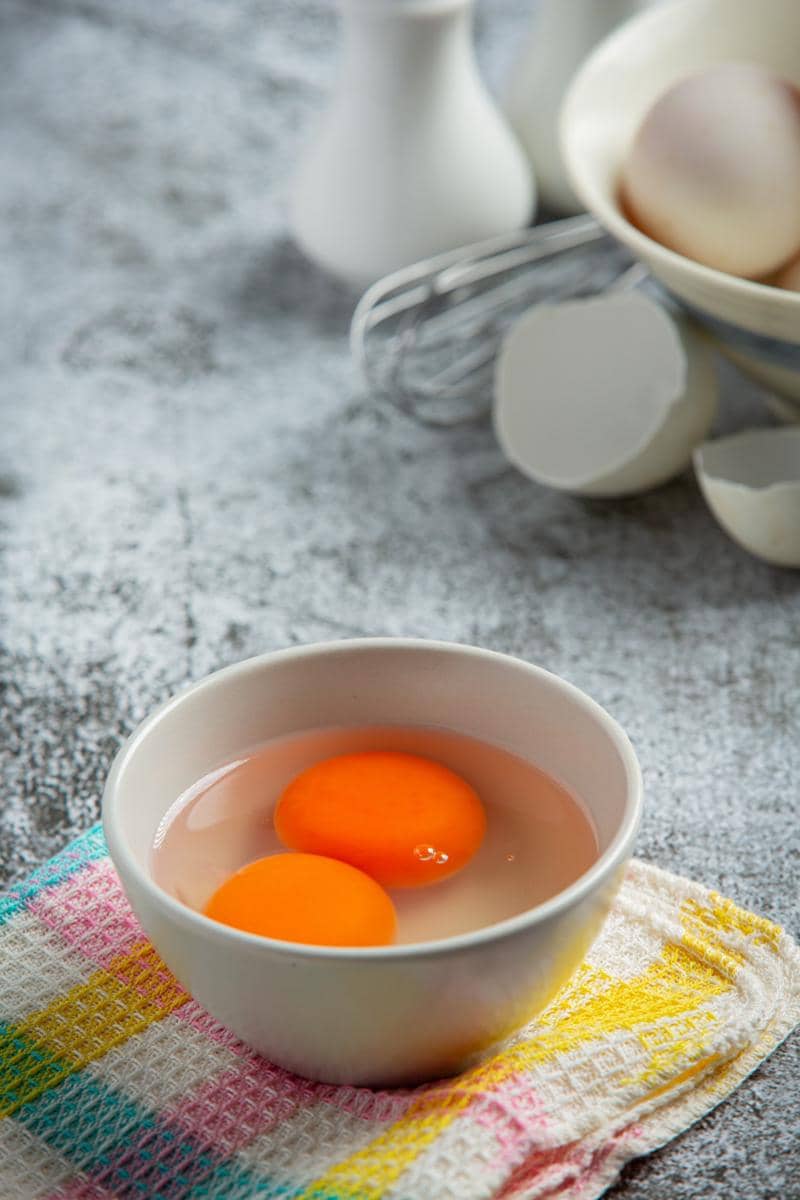 Dua kuning telur omega dalam mangkuk putih di atas kain berwarna pastel dengan latar dapur dan cangkang telur di belakangnya.