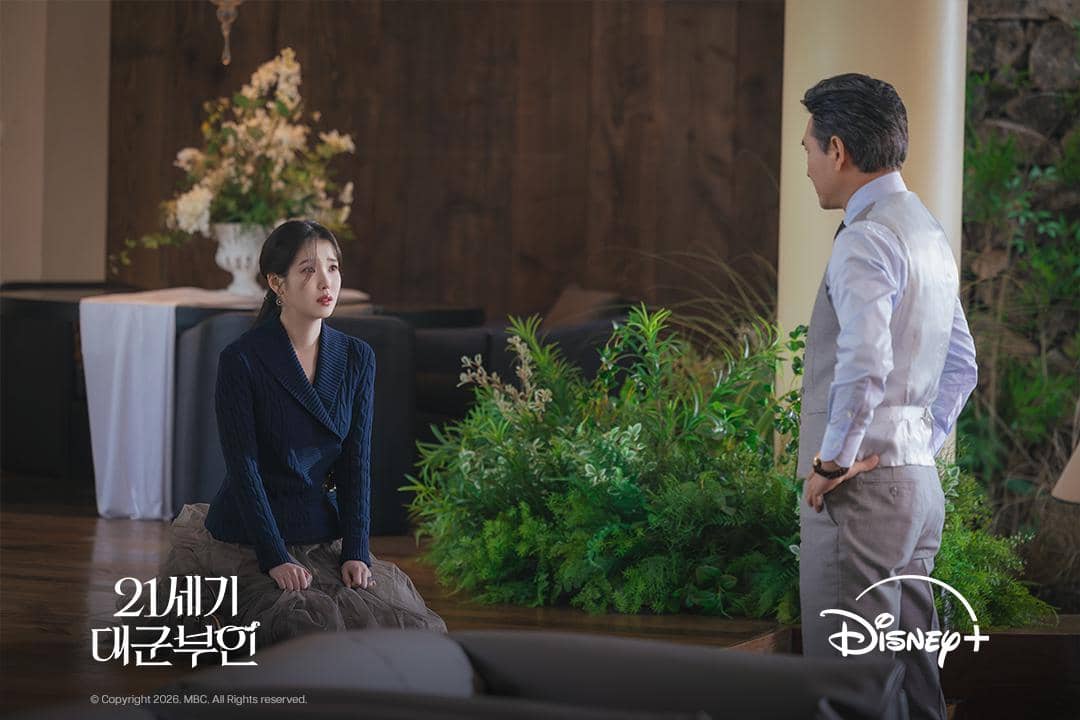Adegan dari drama Korea Perfect Crown menampilkan seorang wanita berlutut di hadapan pria yang berdiri di ruang elegan dengan dekorasi bunga.