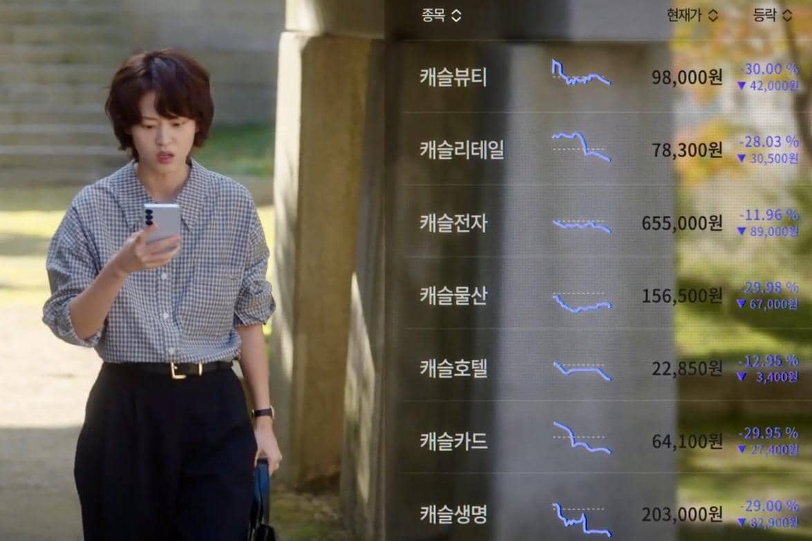 Seorang wanita mengenakan kemeja kotak-kotak memegang ponsel sambil berjalan, dengan tampilan grafik saham dan angka berbahasa Korea di layar.