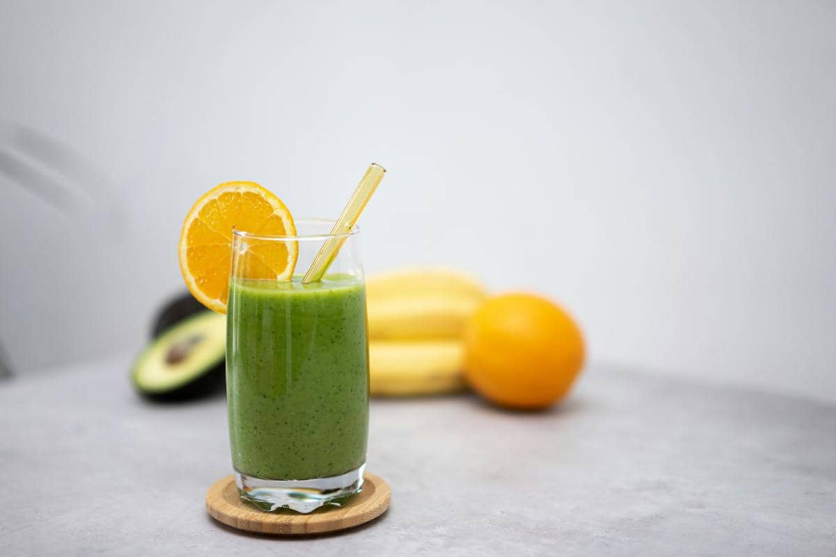 Segelas jus hijau dengan irisan jeruk di tepi gelas dan sedotan kaca, dengan alpukat, pisang, dan jeruk di latar belakang.