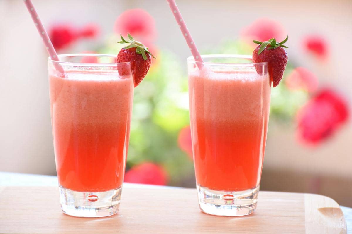 Dua gelas jus stroberi segar berwarna merah muda dengan sedotan dan hiasan buah stroberi di tepi gelas di atas meja kayu.