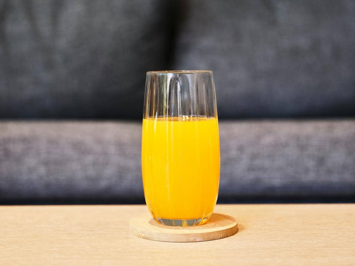 Segelas jus buah berwarna oranye diletakkan di atas tatakan kayu di meja dengan latar belakang sofa abu-abu.