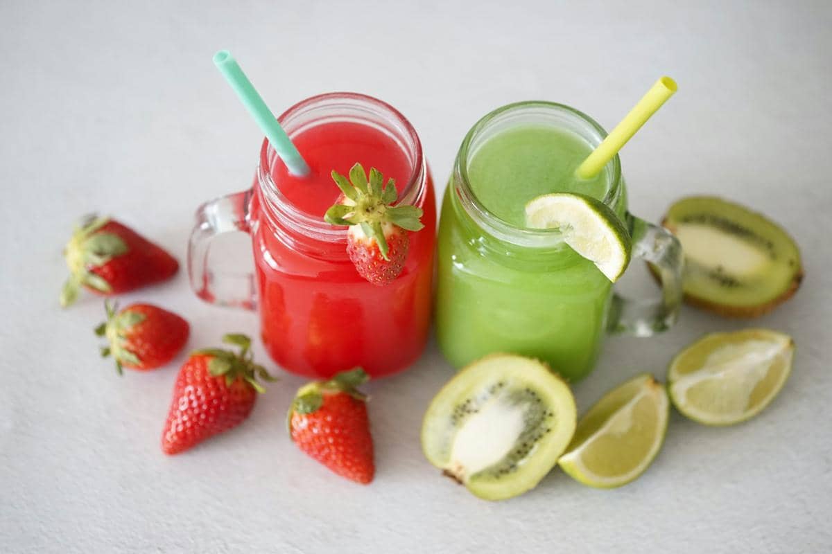Dua gelas jus buah berwarna merah dan hijau dengan sedotan, dihiasi stroberi segar, kiwi, dan irisan lemon di atas meja.