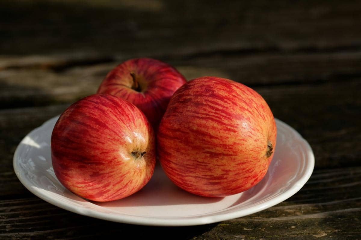 Tiga buah apel merah segar dengan corak garis halus tersusun di atas piring putih di atas permukaan kayu gelap.