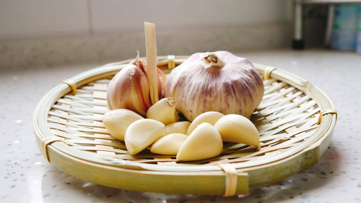 Beberapa siung dan umbi bawang putih segar diletakkan di atas tampah bambu di atas meja dapur yang terang.