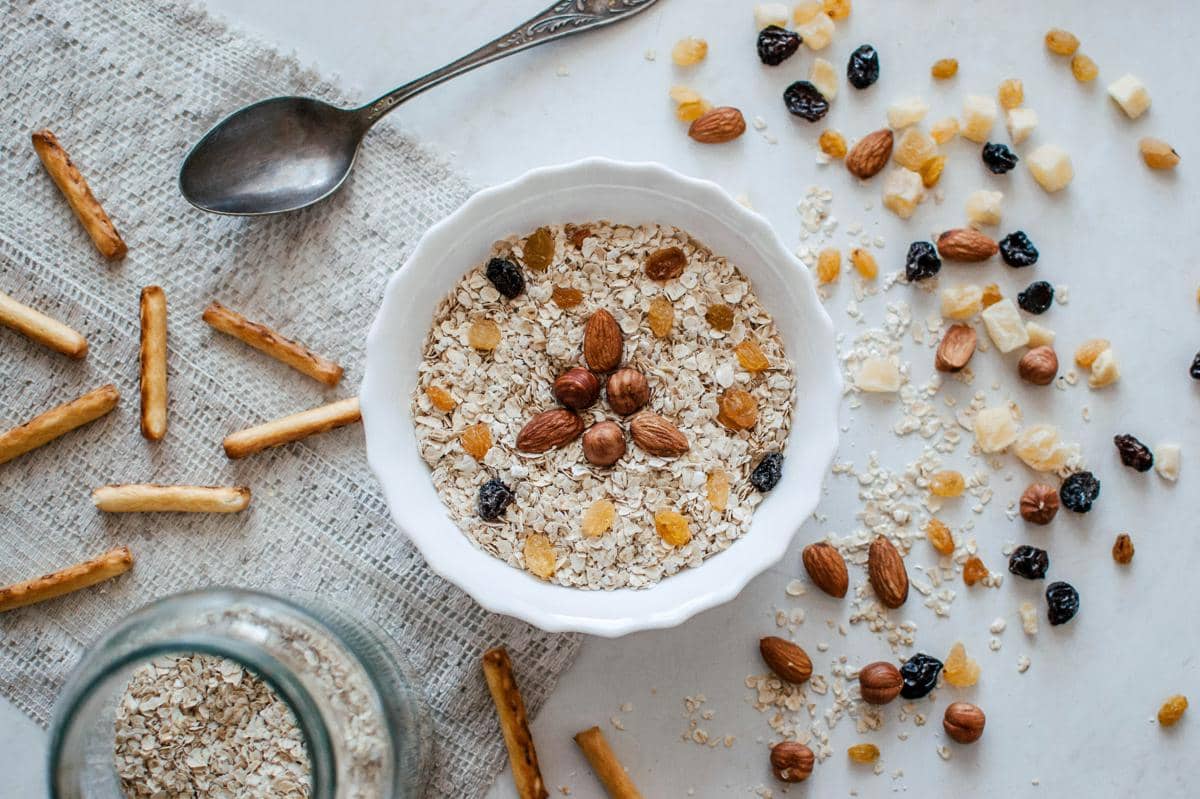 Semangkuk oatmeal dengan kacang almond, kismis, dan potongan buah kering di atas meja bersama sendok dan stik roti.