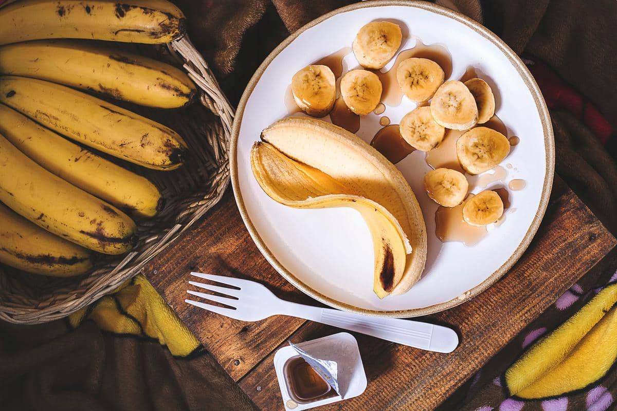 Beberapa pisang utuh dalam keranjang dan potongan pisang di atas piring dengan sirup serta garpu plastik di meja kayu.
