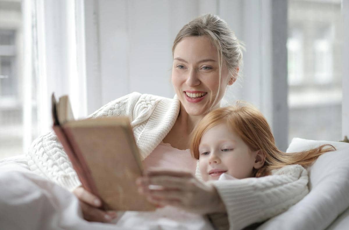 Seorang wanita tersenyum sambil membaca buku cerita bersama anak kecil berambut merah di ruangan terang dekat jendela.