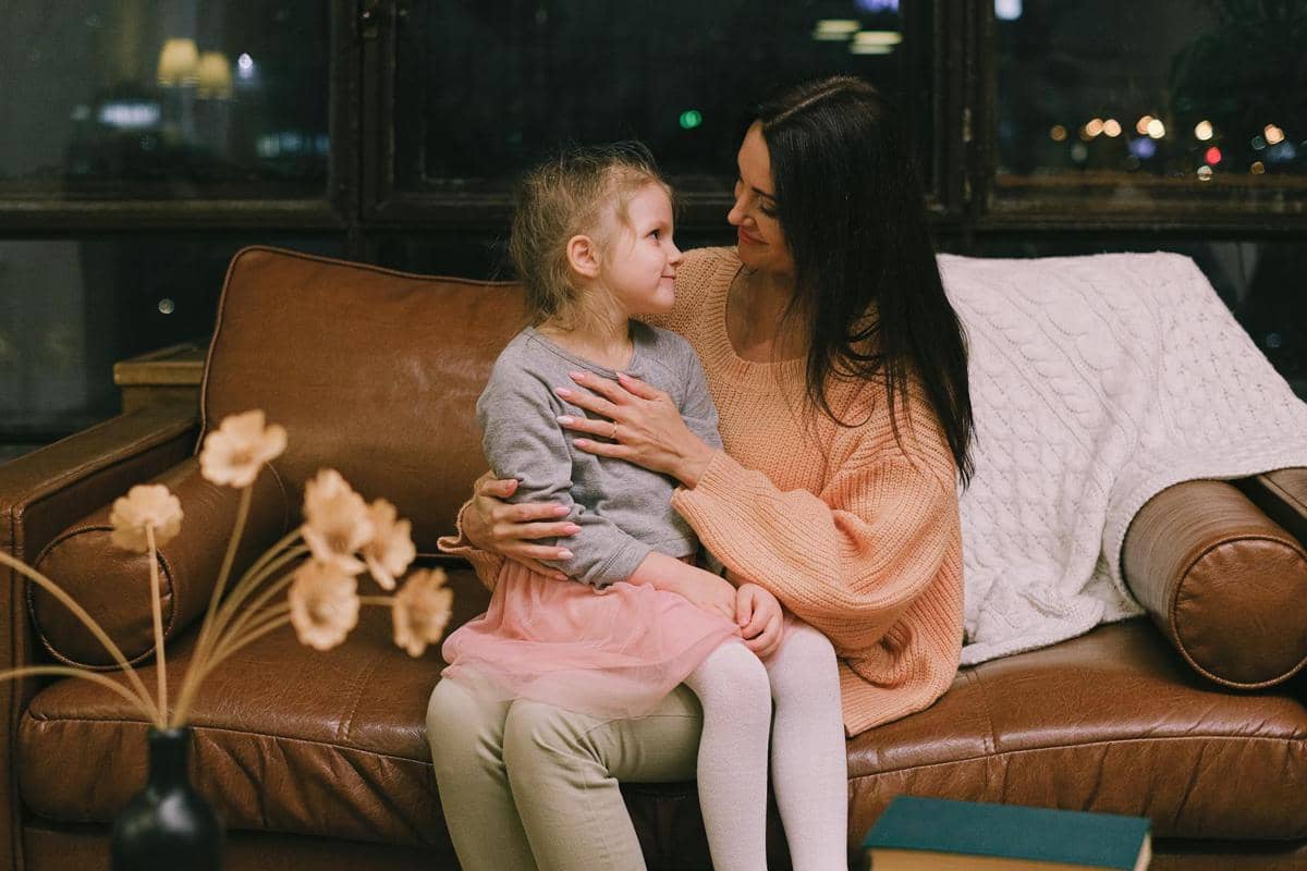 Seorang ibu duduk di sofa sambil menenangkan anak perempuannya yang duduk di pangkuannya, keduanya saling menatap dengan hangat.