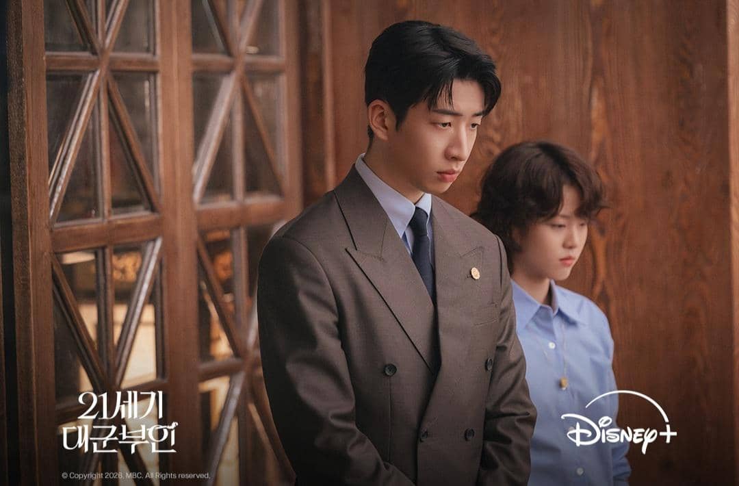 Dua karakter dalam drama Korea Perfect Crown berdiri di ruangan kayu, pria mengenakan setelan jas dan wanita mengenakan kemeja biru.