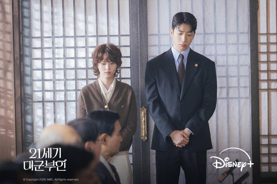 Dua karakter berdiri berdampingan dengan ekspresi serius di ruangan bergaya tradisional Korea dalam cuplikan drama Perfect Crown.