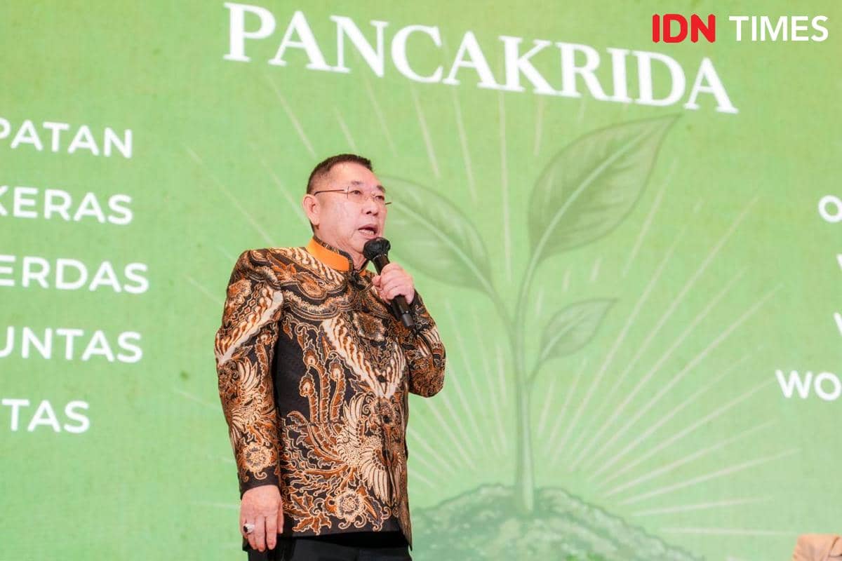Seorang pria mengenakan batik berwarna hitam dan oranye berbicara di atas panggung dengan latar hijau bertuliskan PANCARIDA.