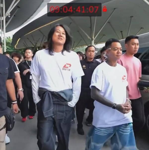 Dua pria berjalan bersama di area luar ruangan dengan beberapa orang di belakang mereka, keduanya mengenakan kaus putih bertuliskan logo merah.