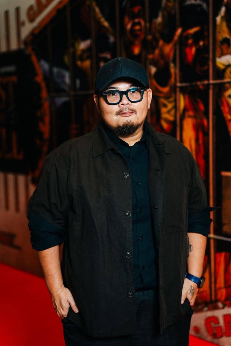 Seorang pria berkacamata mengenakan topi hitam dan pakaian serba hitam berpose di karpet merah dengan latar belakang bergaya seni dinding.