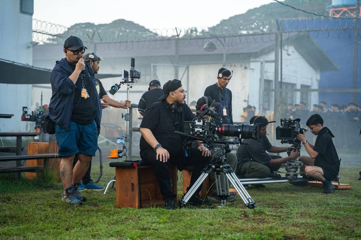 Beberapa kru film sedang bekerja di lokasi syuting luar ruangan dengan peralatan kamera profesional dan suasana pagi berkabut.