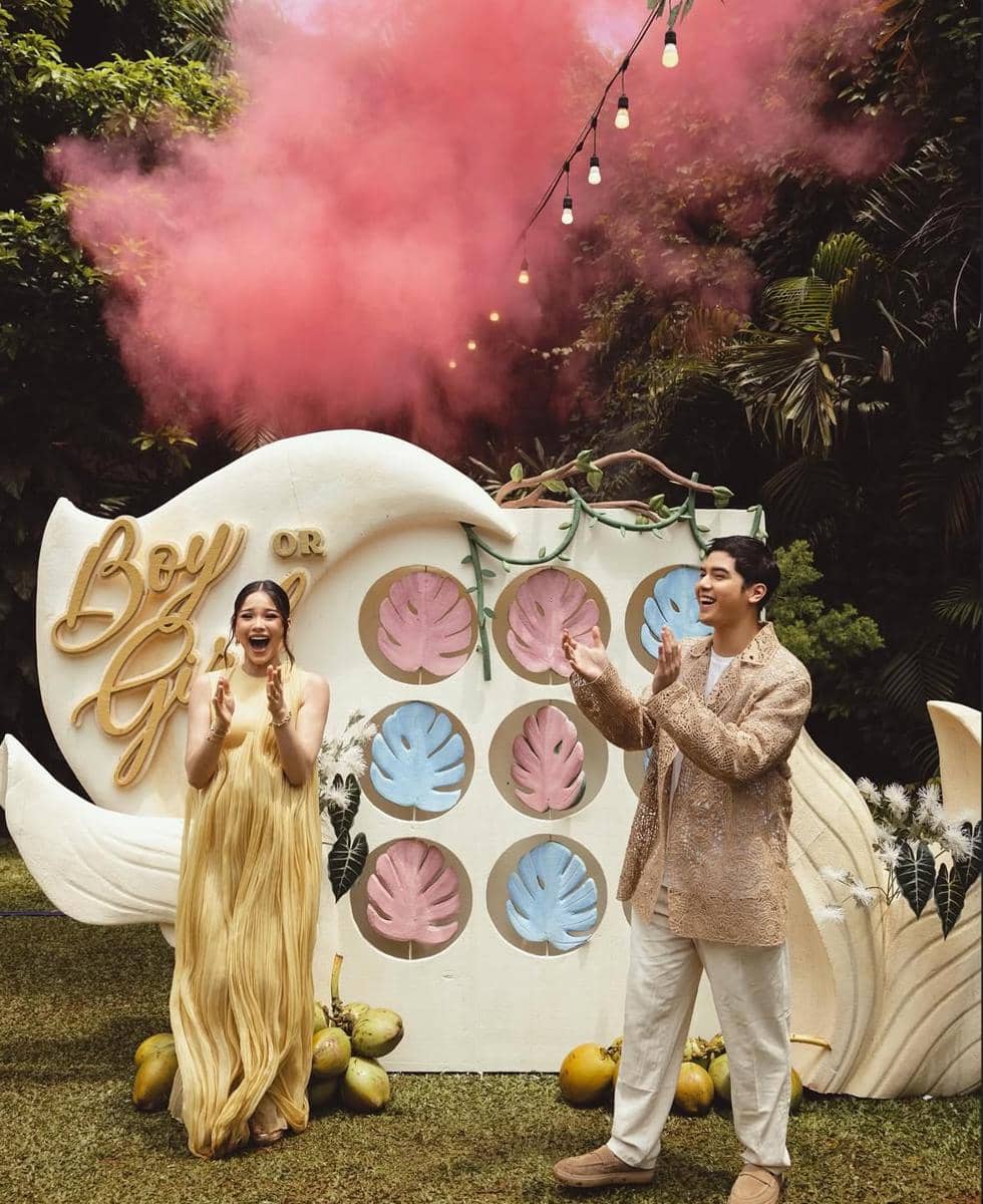 Pasangan tersenyum dan bertepuk tangan di acara gender reveal dengan asap merah muda di taman yang dikelilingi pepohonan hijau.