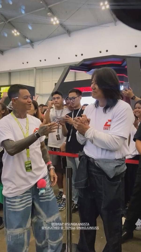 Dua pria mengenakan kaus putih berjabat tangan di acara dalam ruangan dengan banyak penonton di belakang mereka.