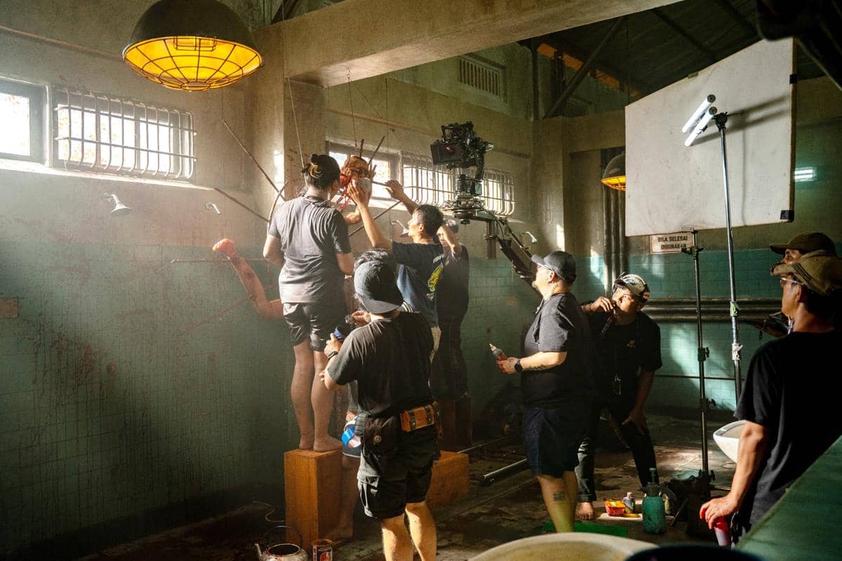 Tim produksi film sedang menyiapkan adegan di lokasi syuting dengan pencahayaan hangat di ruangan berubin hijau dan peralatan kamera profesional.
